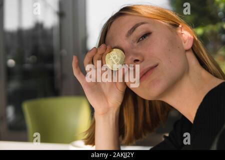 Una bella ragazza è di attaccare una moneta d'oro per il suo occhio. Bitcoins, crypto moneta, il denaro elettronico. Donna seduta al cafe azienda bitcoin nell occhio anteriore Foto Stock