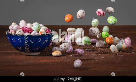 Multi-colore di confetti si è bloccato in movimento in aria accanto a un blu tazza riempita con questi dolci. Le ombre. In legno marrone con sfondo grigio Sfondo. Foto Stock