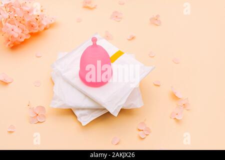 Prodotti per l'igiene femminile - Coppa mestruale e assorbenti igienici sul pallido sfondo arancione con i fiori delle ortensie su di esso. Foto Stock