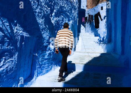 Lone uomo in maglione a strisce che cammina sulle scale con il telefono cellulare in mano nella stretta strada della medina a Chefchaouen (Chaouen), Marocco Foto Stock