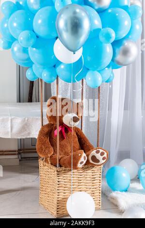 Grande orso di peluche nel palloncino cesto con palloncini blu. Kids arrossire bear giocattolo seduto in un cestello indoor. Bambini decorazione di compleanno Foto Stock