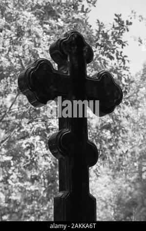 Grave croce di granito nero sullo sfondo del fogliame degli alberi. Il concetto di morte, religione, fede. Immagine in bianco e nero. Foto Stock