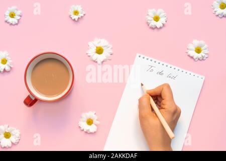 Composizione di fiori con tazza di caffè del mattino e una mano della donna che tiene la penna e scrive 'alla lista di fare ' Foto Stock