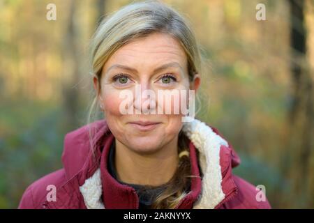 Adulto donna bionda nel suo degli anni quaranta, indossando calda giacca rossa sorridente e guardando la telecamera, mentre in piedi all'aperto nella foresta. Busto ritratto anteriore Foto Stock