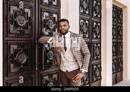 modello maschile africano in un abito marrone che si posa all'esterno, appoggiato sulla parete decorata, porta d'ingresso, elegante lusso costoso business look, ben vestito Foto Stock