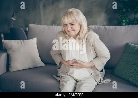 Foto di granny invecchiato dai capelli bianchi nella disperazione che tiene l'addome che soffre il disequilibrio ormonale che siede divano divano divan coperto plaid soggiorno coperto Foto Stock