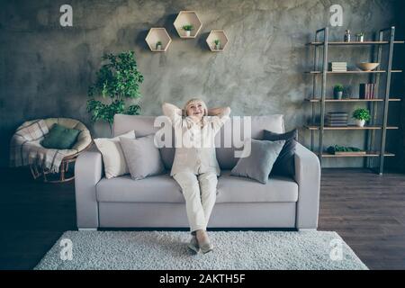 Foto a tutta lunghezza di sorprendente biondo invecchiato granny buon umore mani dietro il soffitto guardando la testa godere di nuovo stile casa design seduta confortevole divano divano divano divano Foto Stock