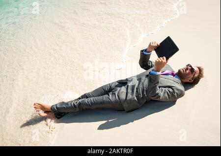 Uomo d'affari rilassato sdraiato in un vestito bagnato utilizzando il suo computer tablet sulla riva di una spiaggia tropicale Foto Stock