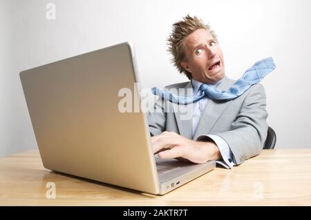 L'uomo d'affari scioccato che apre un file sul suo computer portatile pieno di cattive notizie urlando alla sua scrivania con cravatta volante Foto Stock