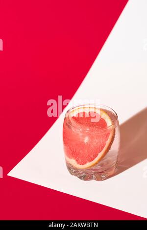 Limonata di pompelmo in un bicchiere con acqua tonica e fetta di pompelmo su sfondo bianco e rosso. Bevande fredde estive. Bevanda detox. Foto Stock