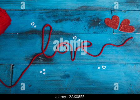 La parola 'amore' con fili di lana rossa e due cuori su sfondo di legno blu. Vista dall'alto San Valentino Foto Stock