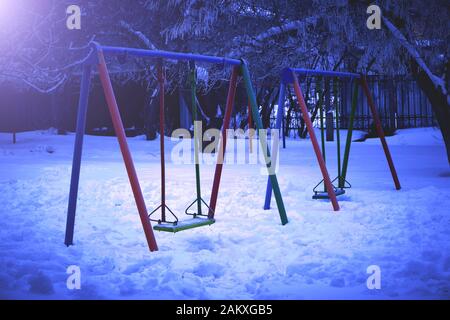 Parco giochi con altalene in inverno di notte alla luce di una lampada da strada. Foto Stock