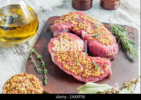 I pezzi di carne sono pronti per la marinata in Dijon senape di grano francese, adagiata su una tavola di argilla con spezie-foglia di baia, pepe, erbe provenzali e olive Foto Stock