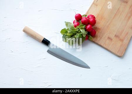 Freschi ravanelli e grande coltello da cucina su sfondo bianco e vecchio tagliere in legno. Copia spazio Foto Stock