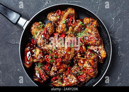 vista dall'alto delle ali di pollo fritte in salsa teriyaki cosparse di semi di sesamo, peperoncino e prezzemolo in una padella su un tavolo di cemento, Foto Stock