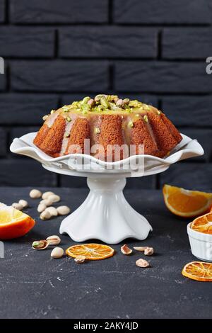 zucca carota bundt torta con smalto arancione, cosparso di pistacchi su un basamento bianco in ceramica con un muro di mattoni sullo sfondo e ingredienti o Foto Stock