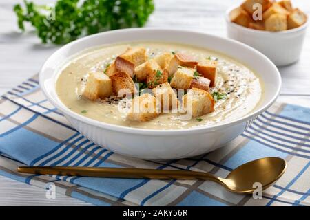Primo piano di una porzione di zuppa di aglio arrosto Asiago Cauliflower sormontata da crostini in una ciotola bianca su un tavolo di legno, vista dall'alto, Foto Stock