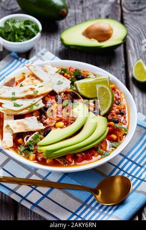 Pollo Taco Soup con fagiolo nero, chicchi di mais, sormontato da tortilla strisce, fette di avocado e calce in ciotola bianca su un rustico tavolo in legno, verticale Foto Stock