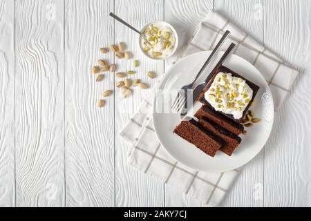 Melassa scura torta di pan di zenzero con panna montata fresca in cima e pistacchi tritati finemente serviti su un piatto bianco su un tavolo di legno, vie orizzontali Foto Stock