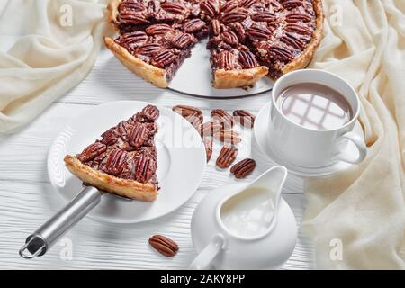 primo piano di deliziosa torta di noci pecan affettata su un piatto bianco e un pezzo servito su un piatto con latte caldo al cioccolato in una tazza, vista dall'alto Foto Stock