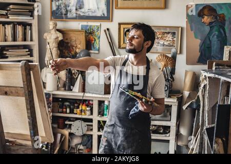 Pittore maschile in studio d'arte all'interno che tiene il pennello davanti controllo prospettiva guardando tela pensiva Foto Stock