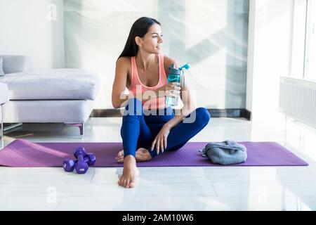 Sensazione di sete. Stanco delle giovani donne in abbigliamento sportivo acqua potabile mentre è seduto sul tappeto di esercizio Foto Stock