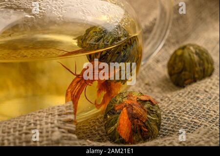 Una sfera di tè del fiore estratto in un teapot di vetro e parecchie sfere di tè del fiore su uno sfondo di tessuto homespun grezzo. Primo piano Foto Stock