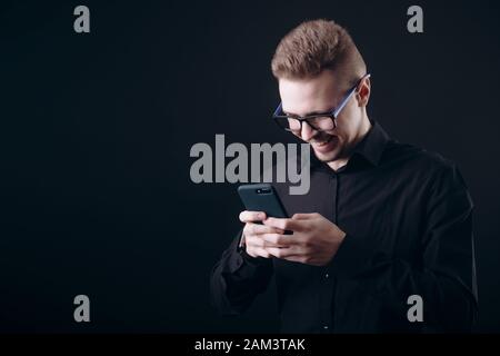 Sorridente barbuto giovane uomo in bicchieri utilizza lo smartphone Foto Stock