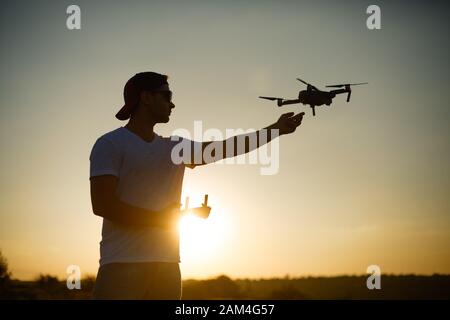 Silhouette di un drone che cattura l'uomo con una mano e telecomando in un'altra mano al tramonto. Sicuro quadcopter atterrare sul palmo del pilota Foto Stock
