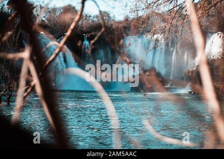 Cascata di Kravice in lontananza, rami di cespuglio sfocati in primo piano. Inverno freddo giorno, acqua blu chiara Foto Stock