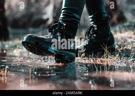 Primo piano di due stivali neri da donna, camminando su una pozza nel campo. Freddo piovoso inverno giorno, maltempo.Prendendo un passo nella giusta direzione Foto Stock