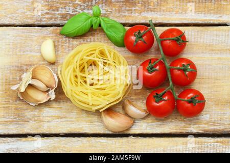 Ingredienti per una pasta italiana perfetta: Tagliatelle, pomodori ciliegini maturi su fusto, spicchi d'aglio e foglie di basilico fresco su una superficie rustica in legno Foto Stock