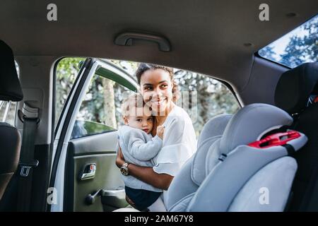 Madre sorridente che mette la figlia del bambino in un sedile dell'automobile. Giovane donna che tiene un bambino sulle mani vicino ad una macchina. Foto Stock