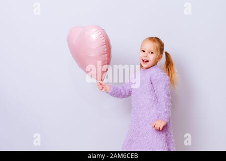 Felice San Valentino cuore giorno. Baby ragazza sorridente con cuore rosa palloncino su sfondo viola chiaro. Foto Stock