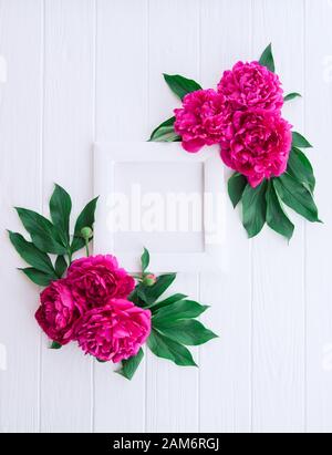 Composizione elegante con cornice fotografica con tela bianca e fiori rosa viola di peonia con foglie verdi sullo sfondo bianco di legno. Scheda verticale. Foto Stock