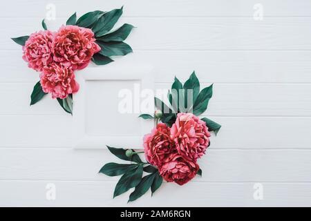 Elegante composizione con cornice fotografica con tela bianca e fiori rosa rosa rosa peonia con foglie verdi sullo sfondo bianco di legno. Tono di colore disattivato Foto Stock