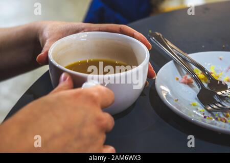mani che tengono una tazza mezza bevuta di tè con il piatto vicino vicino con le briciole della torta sopra Foto Stock