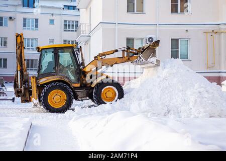 Attrezzatura per la rimozione della neve su strada in inverno. L'escavatore giallo rimuove un'enorme montagna di neve. Spazzare la neve sulla strada in città in una tempesta di neve. Foto Stock