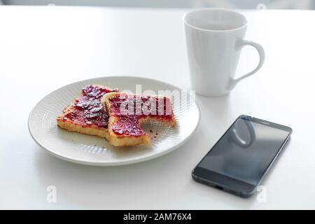 La colazione è a tavola. Una tazza di caffè e toast con marmellata di ciliegie accanto al telefono. Foto Stock