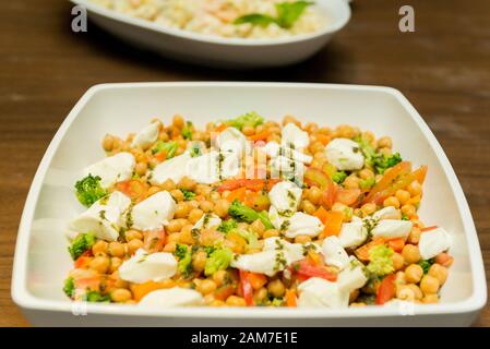 Primo piano di insalata di Chickpea con pomodori, broccoli e formaggio di bufalo su un piatto di ceramica Foursquare su un tavolo di legno. Cibo sano e gustoso. Selezione Foto Stock