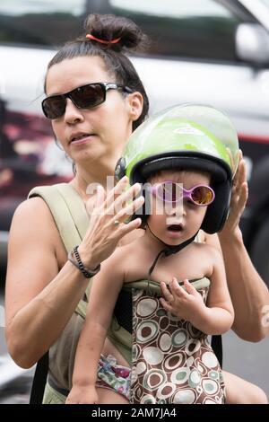 Giovane madre turistica che mette un casco verde di scooter di calce sul suo vetro di sole rosa che indossa cute, figlia del toddler prima di ottenere sullo scooter. Foto Stock