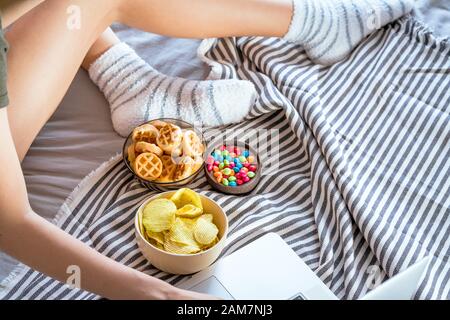 Ragazza lavora a un computer in un letto e mangia fast food. Cibo non sano: Patatine fritte, cracker, caramelle, waffle. Cibo spazzatura, concetto. Foto Stock