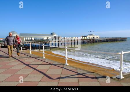 Coppia passeggiando lungo il lungomare di Hastings di fronte al molo; sotto cieli nudless, in una soleggiata giornata invernale, East Sussex, Regno Unito Foto Stock