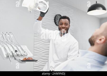 Dentista maschio intelligente in uniforme bianca che lavora e cura il mal di denti in clinica. Uomo africano professionista che guarda la macchina fotografica e sorridente mentre il cliente sdraiato sulla sedia. Concetto di procedure dentali. Foto Stock