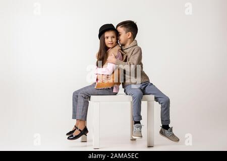 Ragazza e ragazzo carino ed elegante in abiti alla moda seduti insieme in studio. Bambino ragazzo abbraccia la ragazza e le racconta una storia divertente. Bambini fash Foto Stock