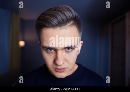 Pericoloso sguardo di un giovane depresso in un ambiente cupo di una stanza blu profondo. Un maschio infastidito di 20 anni sembra sullenly. Ragazzo arrabbiato guarda da b Foto Stock