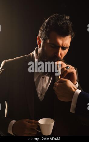 Uomini luci di mano busy businessmans sigaro per fumare. Pranzo d'affari e concetto di pausa di cinque minuti. L'uomo con la barba tiene sigaro e caffè su sfondo grigio. Il ragazzo in abito classico ha pausa caffè al mattino. Foto Stock