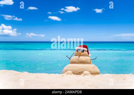 Sorridente pupazzo di neve sabbiosa con cappello rosso santa sulla spiaggia caraibica con cielo bellissimo . Concetto di vacanza per Capodanno e carte di Natale Foto Stock