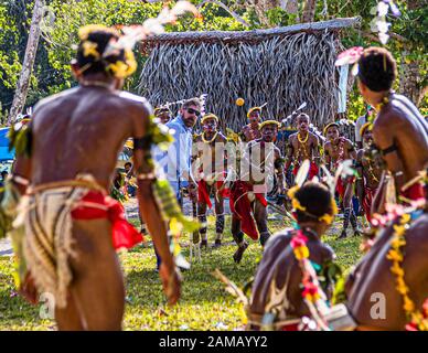 Un passeggero della nave da spedizione True North lancia la palla arancione durante una partita di cricket in stile Isole Trobriand a Kwebwaga, Isole Trobriand, Papua Nuova Guinea, un ballerino in costume corre verso di lui, uomini in costume stanno in piedi intorno al wicket davanti a una capanna con tetto di foglie di palma. Foto Stock