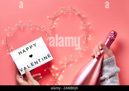 San Valentino piatto, vista dall'alto su sfondo rosa. Mani che tengono lightbox con testo 'Happy Valentine' e bottiglia di champagne rosa. Ghirlanda leggera in hea Foto Stock